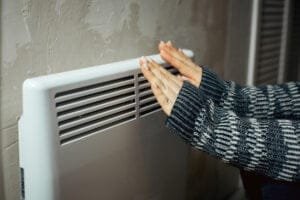 a woman warms her hands at the radiator in a cold house, problems with heating, heating the room with an electric convector